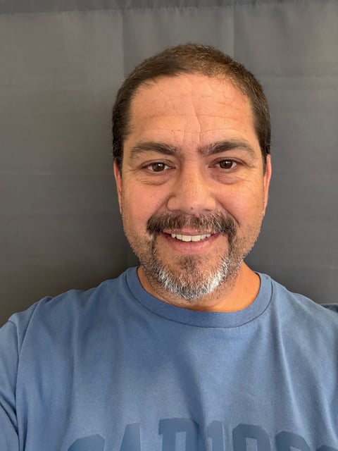 Portrait of a middle-aged man with brown hair and beard wearing a blue shirt, smiling at camera against gray background